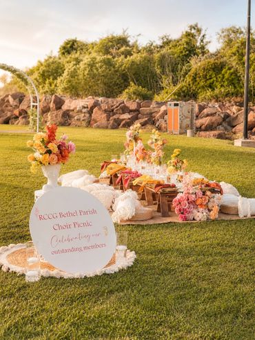 Outdoor picnic setup with flowers and sign celebrating choir members.
