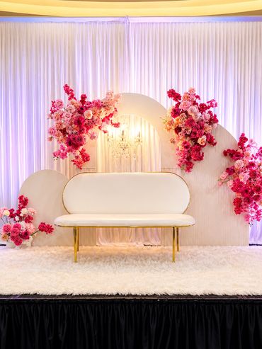 Elegant white sofa with golden legs surrounded by vibrant pink floral arrangements on a stage.