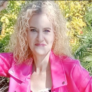 Woman with curly blonde hair wearing a bright pink jacket outdoors.