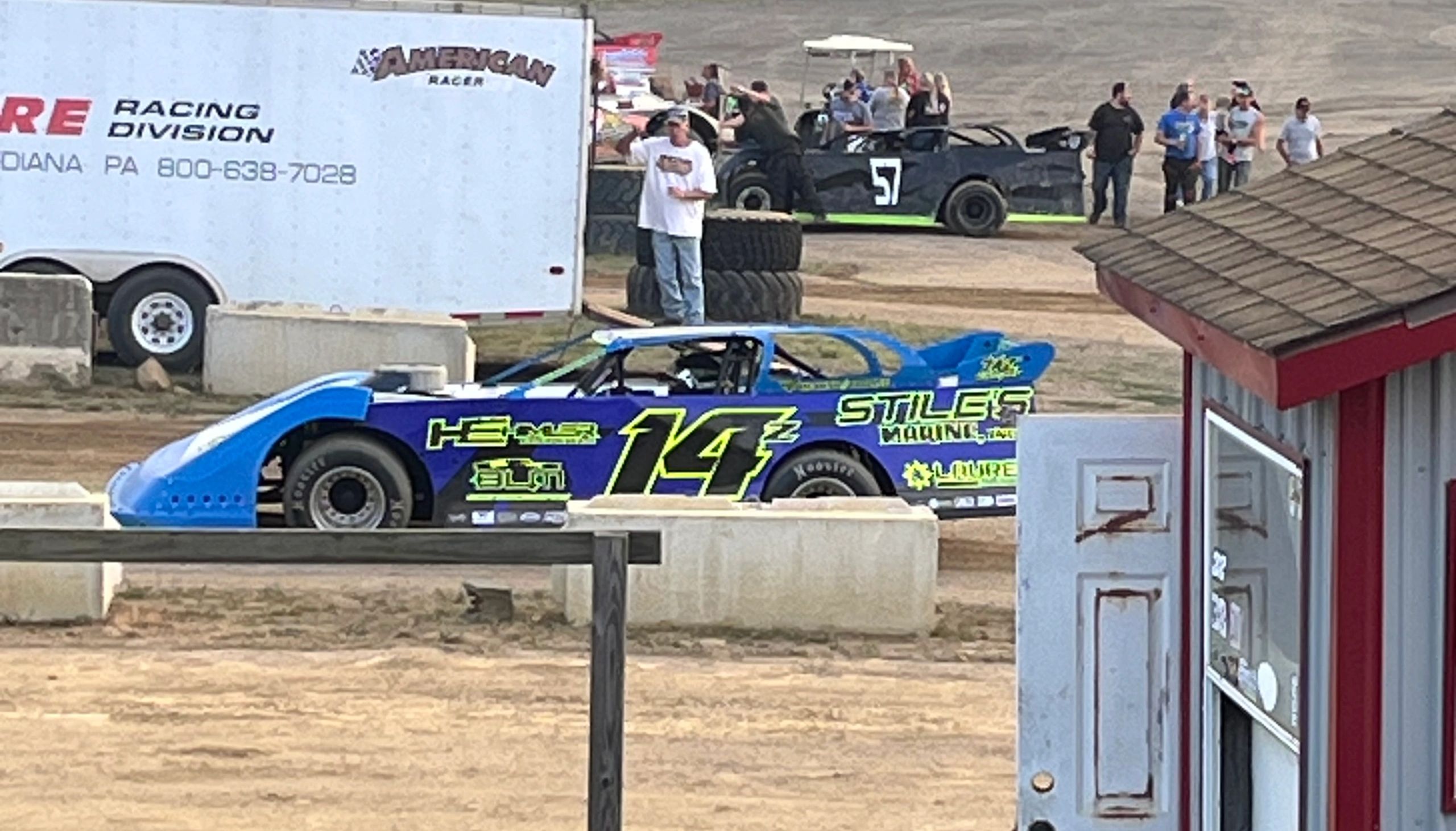Race cars and people at a dirt track racing event with a blue and yellow car in the foreground.