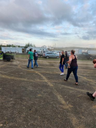 People walking at an outdoor event with vehicles and trailers in the background.