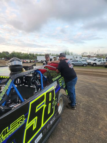 Two men working on a black and neon green race car numbered 58 on a dirt track.