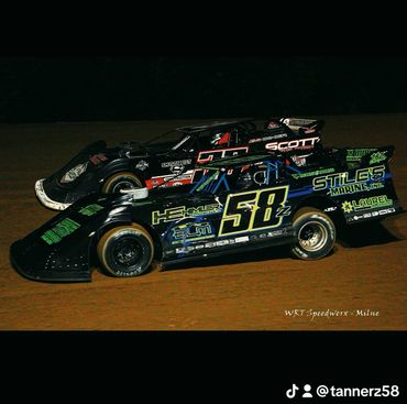 Two dirt late model race cars competing on a dirt track at night.