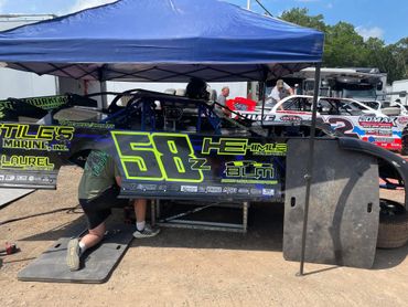 Mechanic working on a race car under a blue canopy in a dirt pit area.