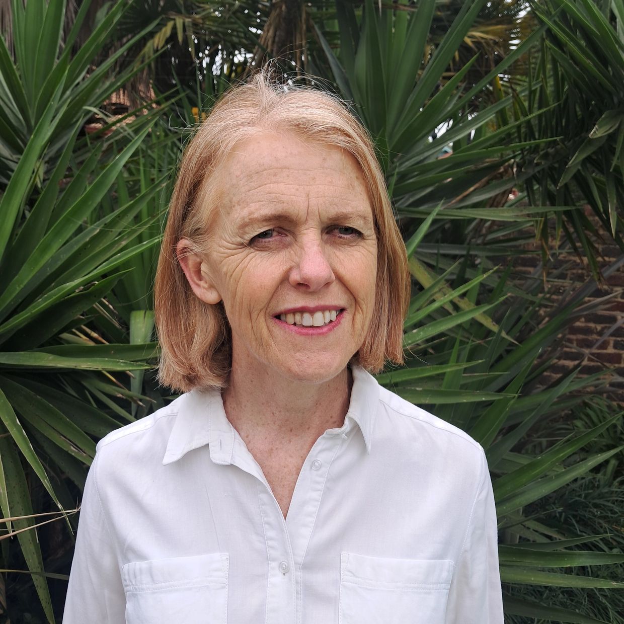 A smiling woman in a white shirt stands in front of green foliage.
