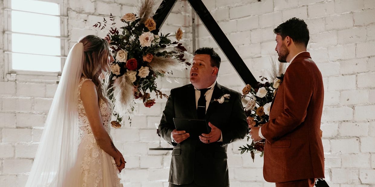 DFW wedding officiant leading a couple's ceremony in front of a floral arch at an industrial-style brick venue