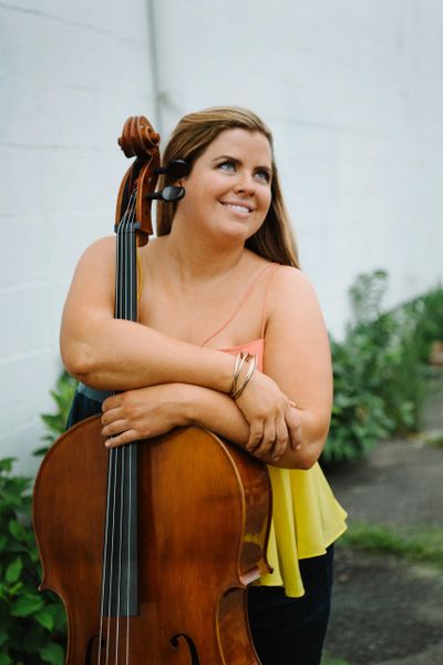 Woman smiling while holding a cello outdoors.