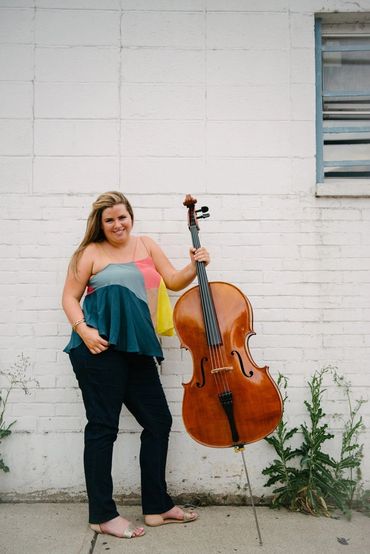 A woman stands holding a cello against a white brick wall.