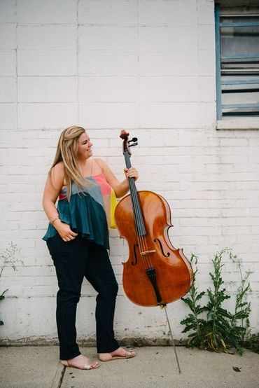 Woman standing outdoors holding a cello against a white brick wall.
