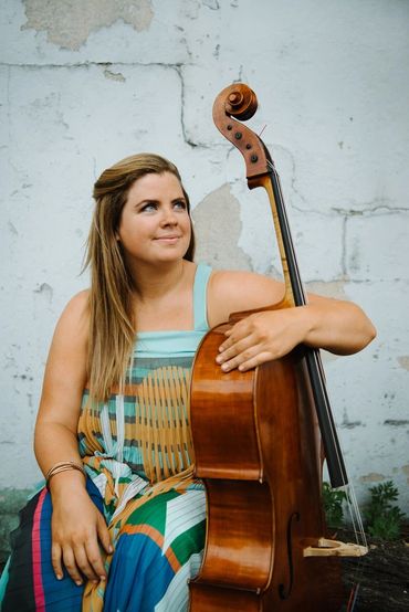 A woman in a colorful dress holding a cello against a weathered wall.