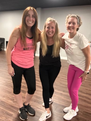 Three women smiling in workout clothes indoors.