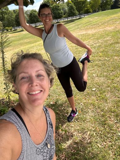 Two women smiling outdoors, stretching in a park.
