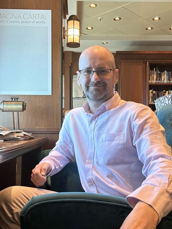 Matthew Clements-Wheeler in smart casual attire seated in a library smiling at the camera
