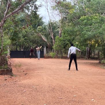 Three people playing cricket on a dirt ground surrounded by trees.