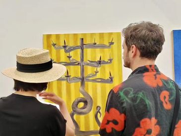Two people observe a modern art piece featuring a yellow background and abstract shapes.
