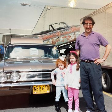 Man and two young girls pose in front of a classic car with a rusty vehicle on a trailer behind them.