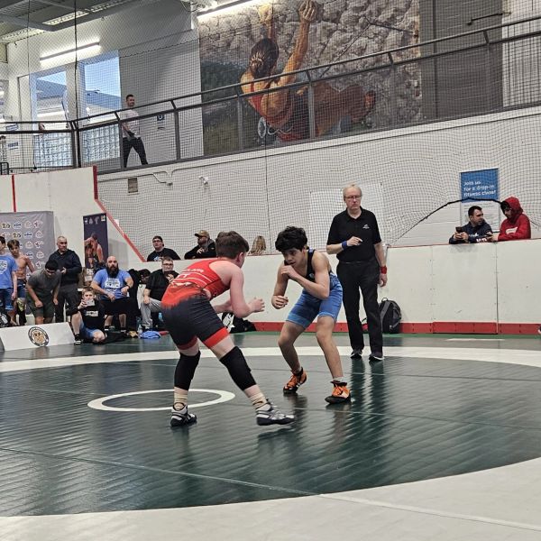 Two young wrestlers facing off on a mat during a match with spectators and a referee nearby.