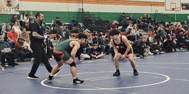 Two wrestlers in a gymnasium preparing to start a match with a referee nearby.