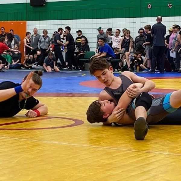 Two wrestlers compete on a yellow mat with a referee closely observing.