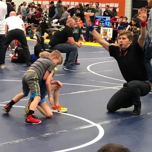 Two young boys wrestling on a mat with a referee signaling a move.