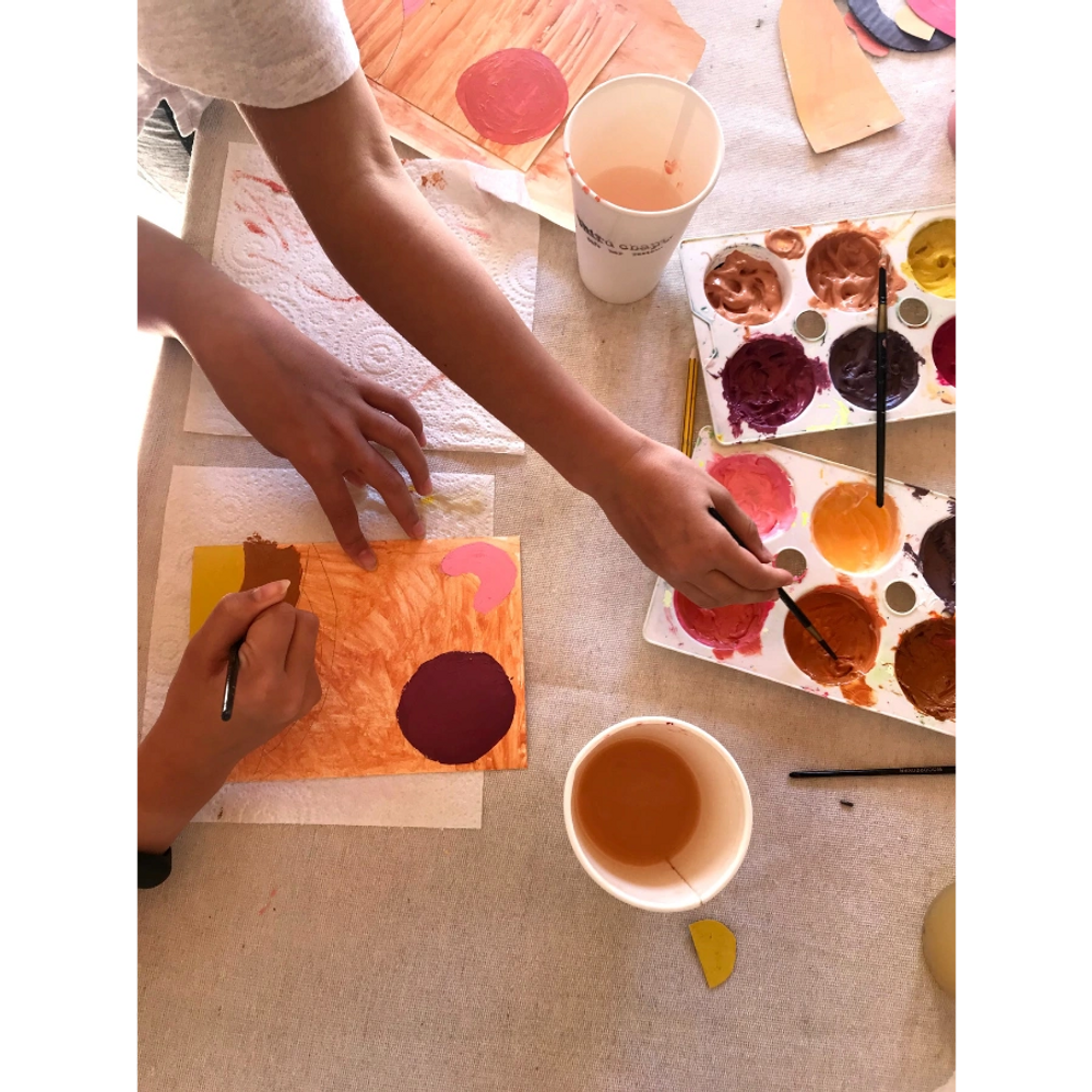 A table is filled with warm coloured paints. Arms stretch to reach paint palettes. 