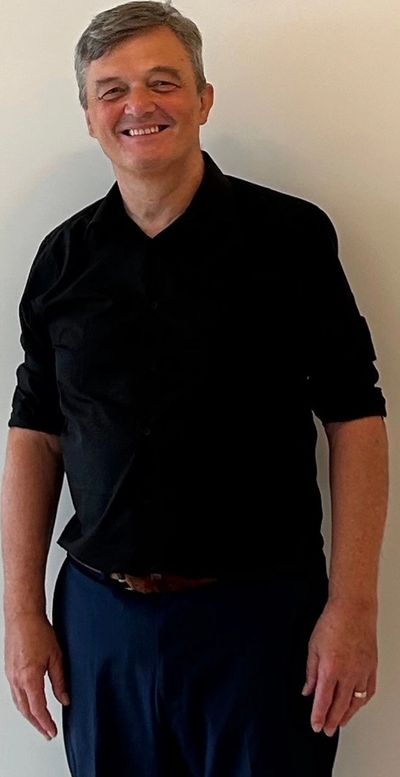 Man smiling in black shirt and blue pants against a plain background.