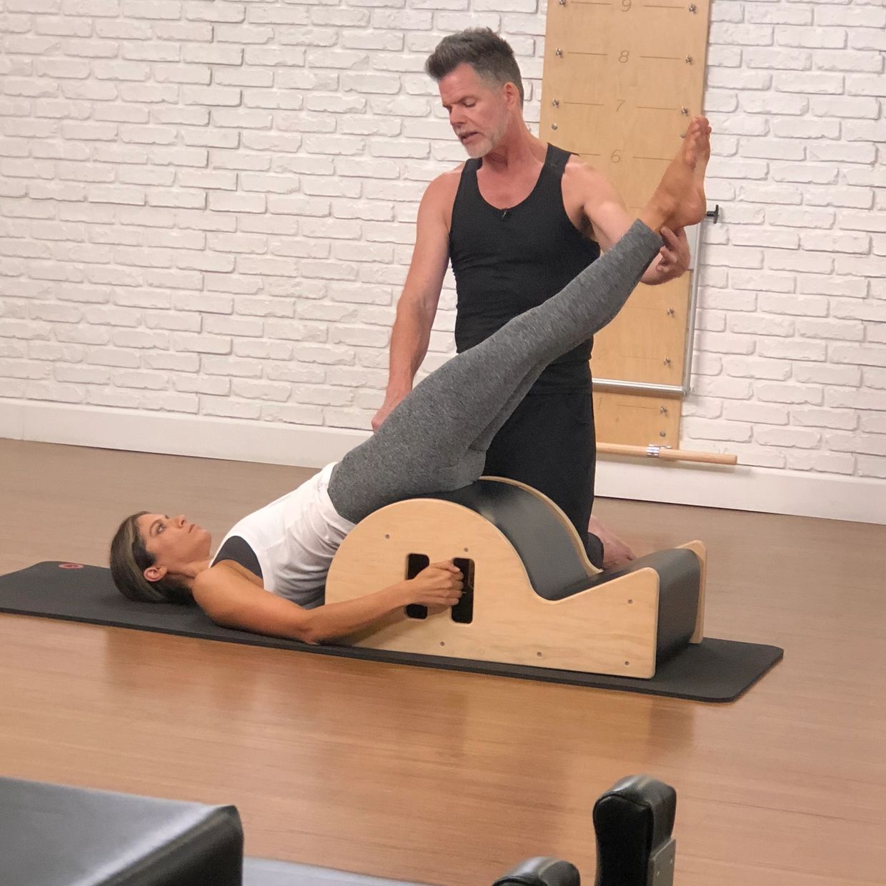Woman performing Pilates exercise with instructor assistance on a wooden arc barrel.