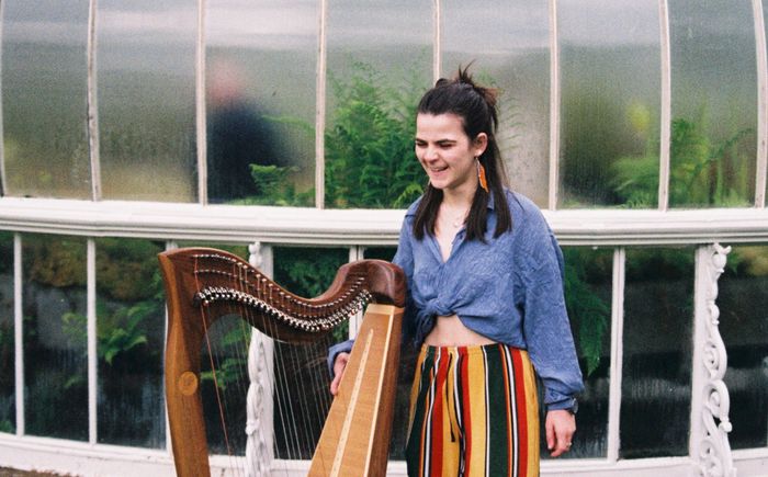 Grace smiling and looking at her harp standing in front of a greenhouse, wearing funky trousers.