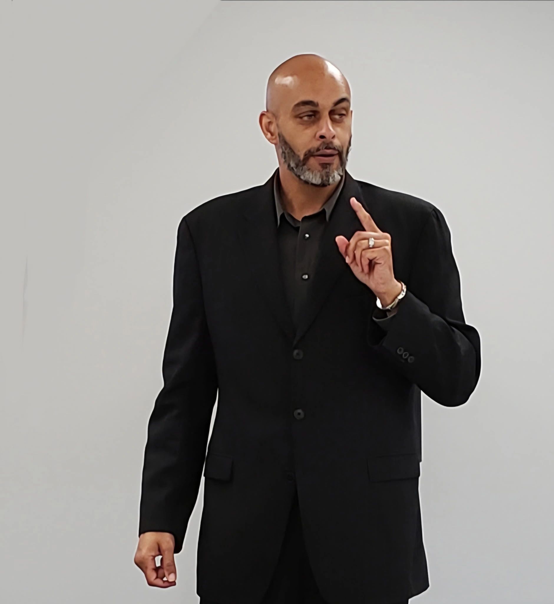 Man in black suit gesturing with raised finger while speaking.