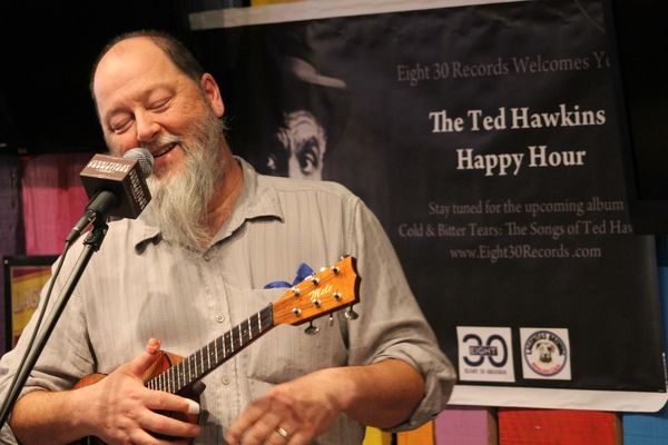 Bearded man playing ukulele and smiling into microphone.
