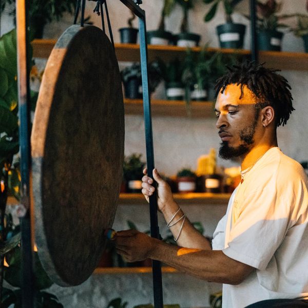 Man playing a large gong in a cozy plant-filled room.