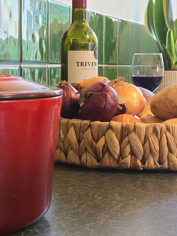 A kitchen counter with onions, a potato, a wine bottle, and a glass of red wine.