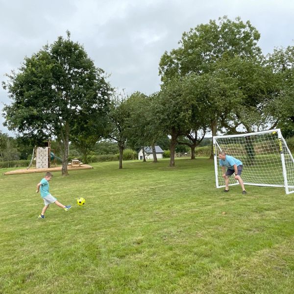 Young boy and his dad playing football together in the orchard. Wooden play structure in the backgro