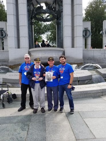 Four men in blue shirts at a memorial, with two veterans holding a photo.