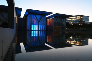 Modern buildings with blue and orange lights reflecting on water at dusk.