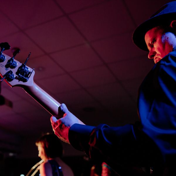 Musician playing guitar on stage with singer in background.