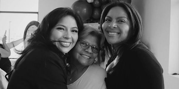 Three women smiling and hugging joyfully indoors.