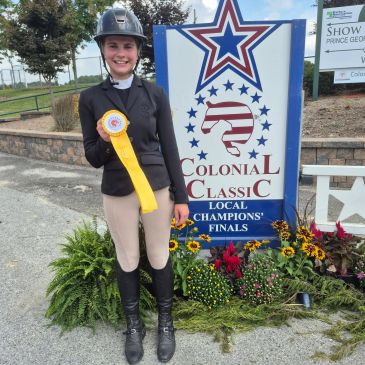 Girl in equestrian attire holding a yellow ribbon at a horse show.