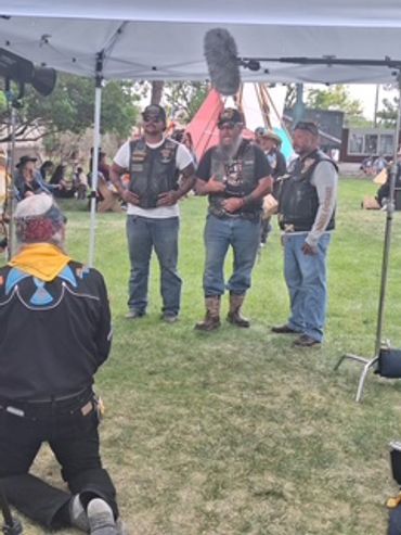 Three men in biker vests standing under a canopy in a park setting.