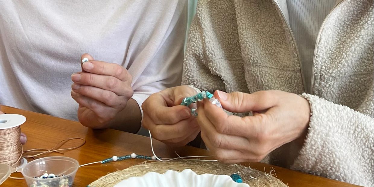 Two people making beaded bracelets together at a wooden table.