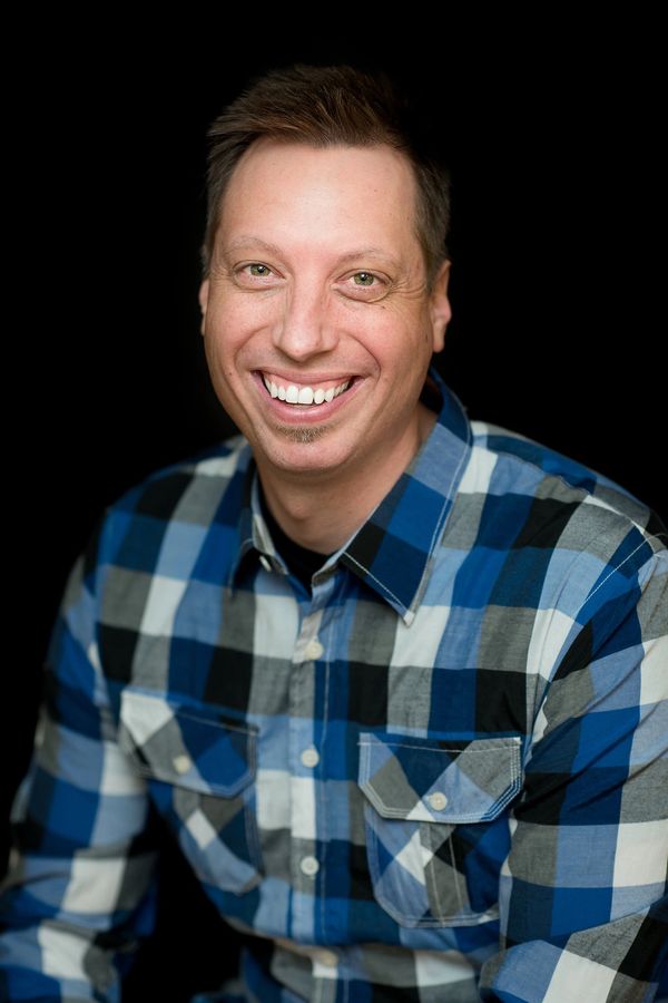 Smiling man in a blue and black checkered shirt against a black background.