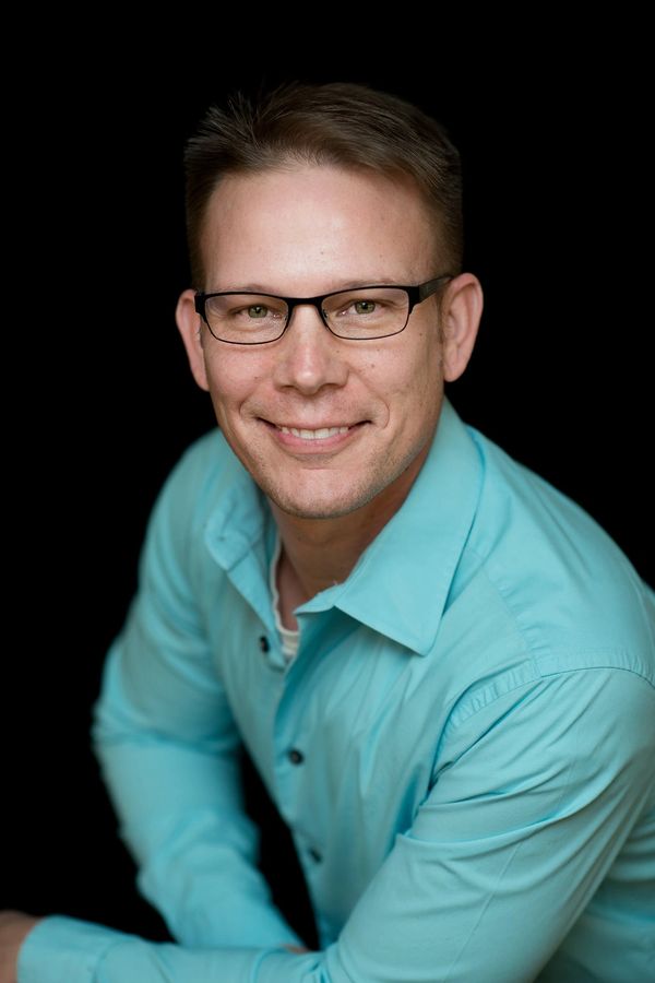 Smiling man with glasses in a turquoise shirt against black background.