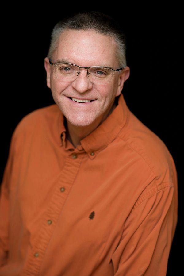 Smiling man in glasses wearing an orange shirt against a black background.
