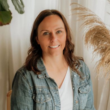 Smiling woman in a denim jacket posing indoors with plants in the background.