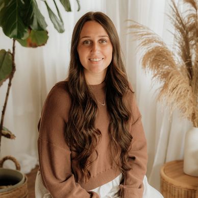 Young woman with long brown hair sitting indoors, smiling gently.