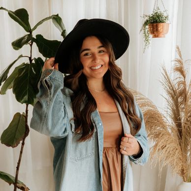 A smiling woman in a black hat and denim jacket poses indoors surrounded by plants.