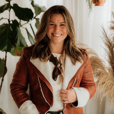 Smiling woman in a stylish brown shearling jacket poses indoors with plants in the background.