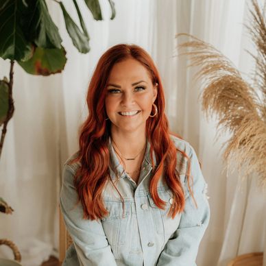 Smiling woman with red hair wearing a distressed denim jacket, sitting on a chair indoors.