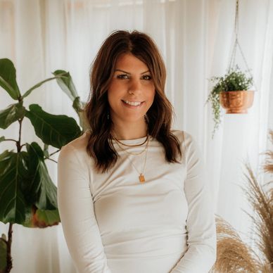 Young woman with brown hair in a white top and ripped jeans smiling indoors.