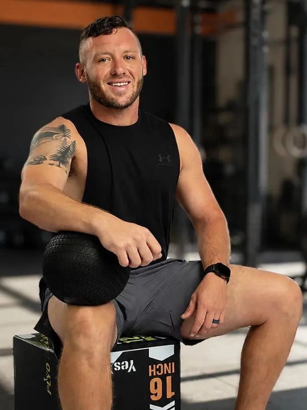 Smiling man in gym attire sitting with a black medicine ball.
